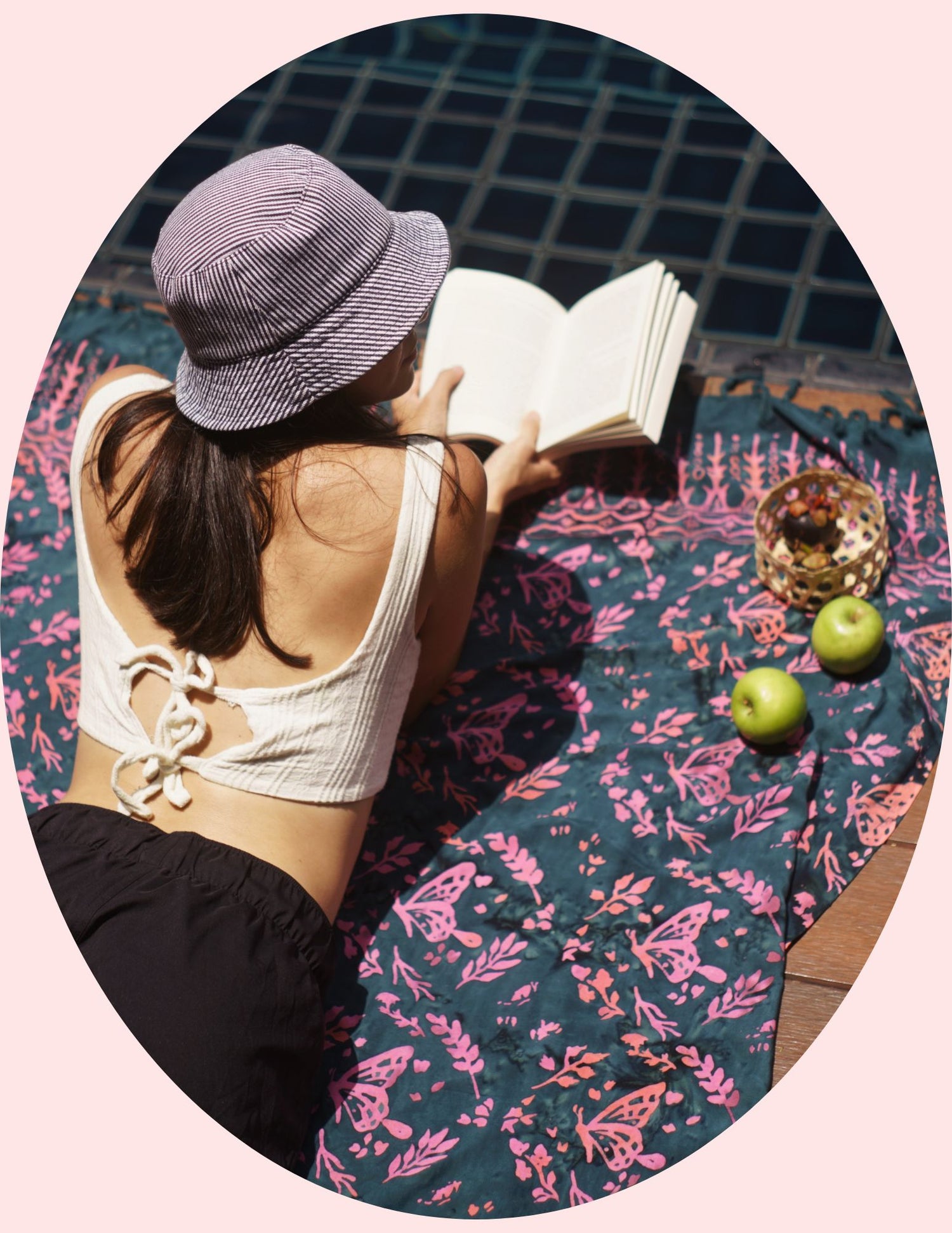 The Butterfly - Dark Slate sarong from YUMI & KORA is used as a pool towel. A woman reads a book on top of the sarong by the pool