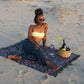 The Butterfly - Dark Slate sarong from YUMI & KORA is used as a beach towel. A woman sits on the sarong at the beach with a picnic basket