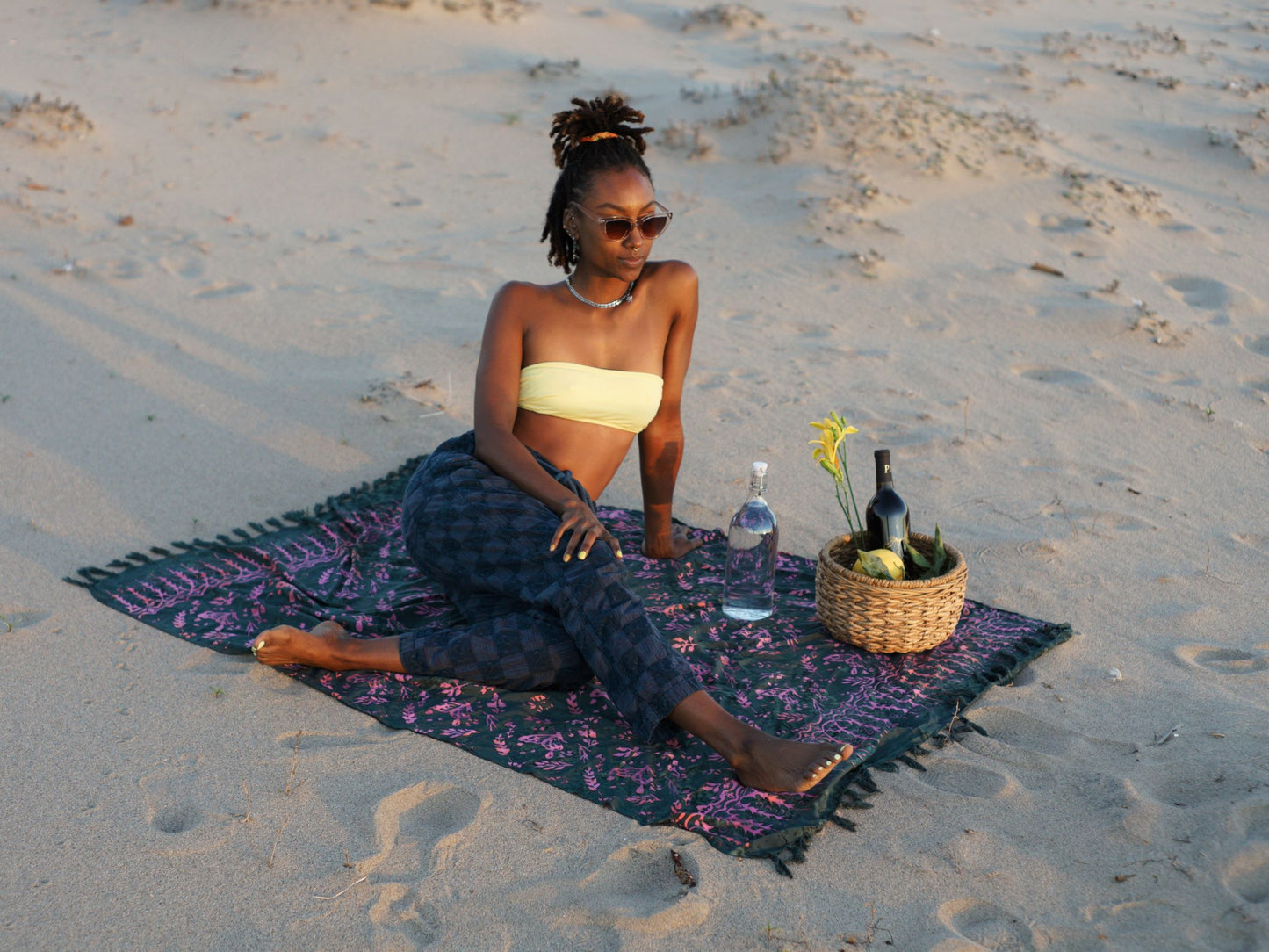 The Butterfly - Dark Slate sarong from YUMI & KORA is used as a beach towel. A woman sits on the sarong at the beach with a picnic basket