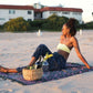 The Butterfly - Dark Slate sarong from YUMI & KORA is used as a beach towel. A woman sits on the sarong at the beach with a picnic basket