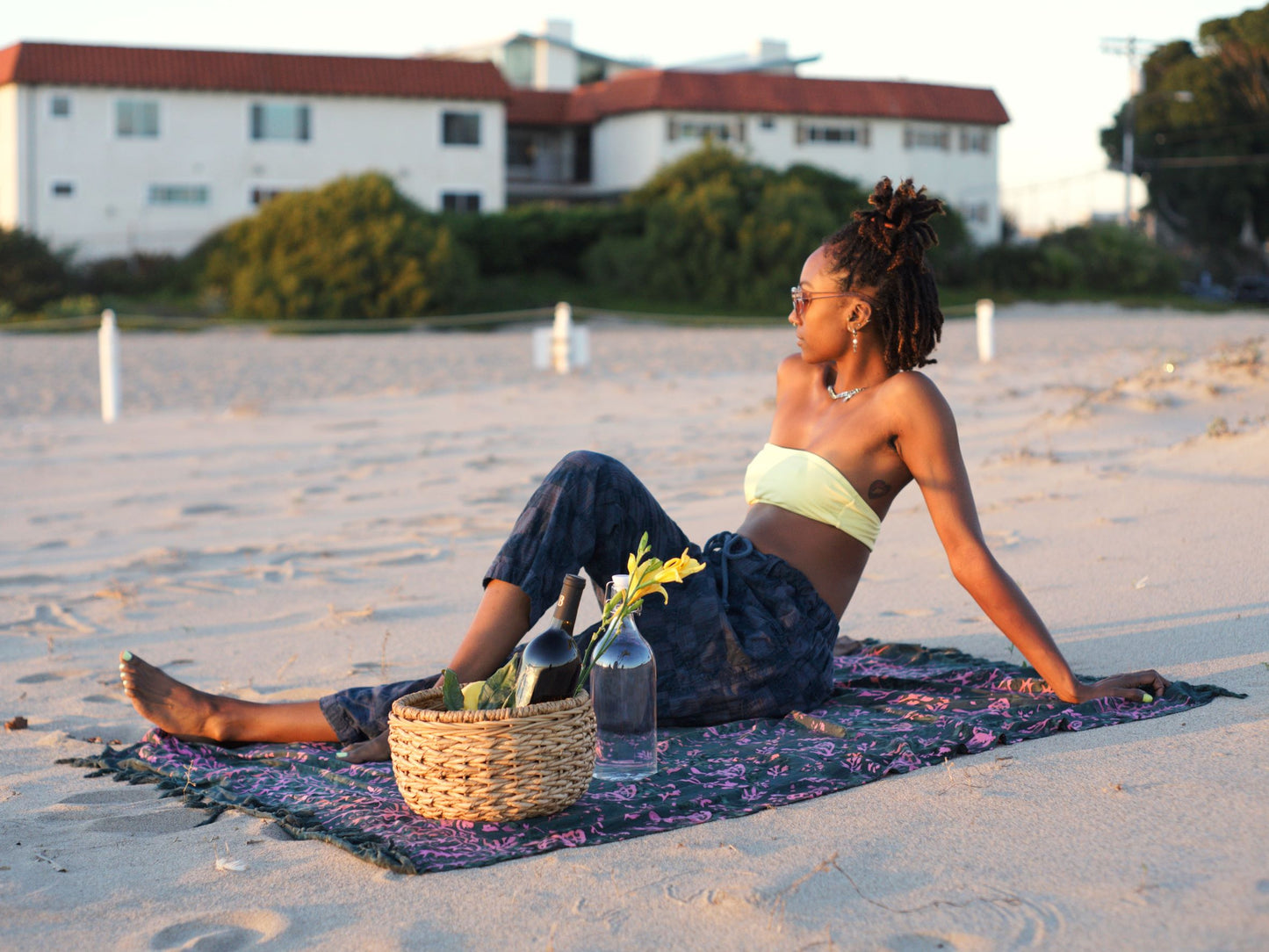 The Butterfly - Dark Slate sarong from YUMI & KORA is used as a beach towel. A woman sits on the sarong at the beach with a picnic basket