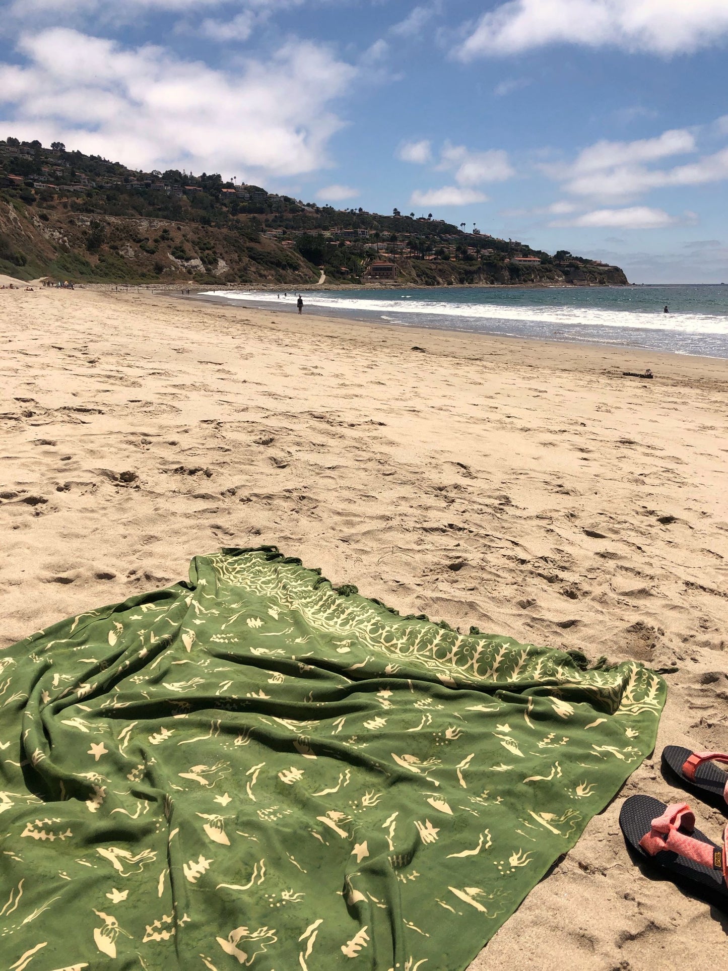 The Duck Dive - Olive Green sarong from YUMI & KORA sits on the sand at the beach. Our sarongs can be used as a beach towel or as a pool towel