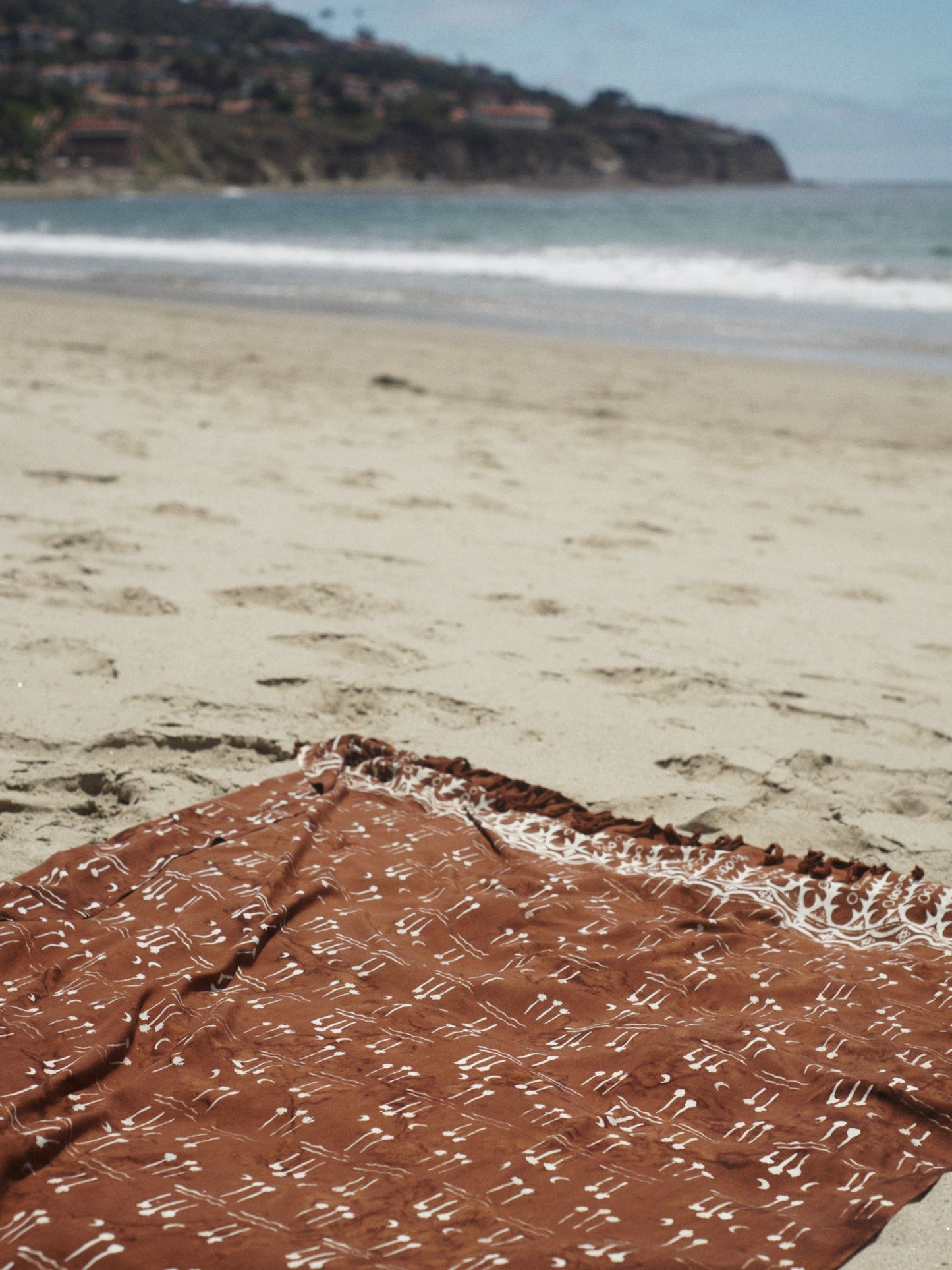 The Hin Kong Bay - Brown sarong from YUMI & KORA sits on the sand at the beach. Our sarongs can be used as a pool towel or as a beach towel