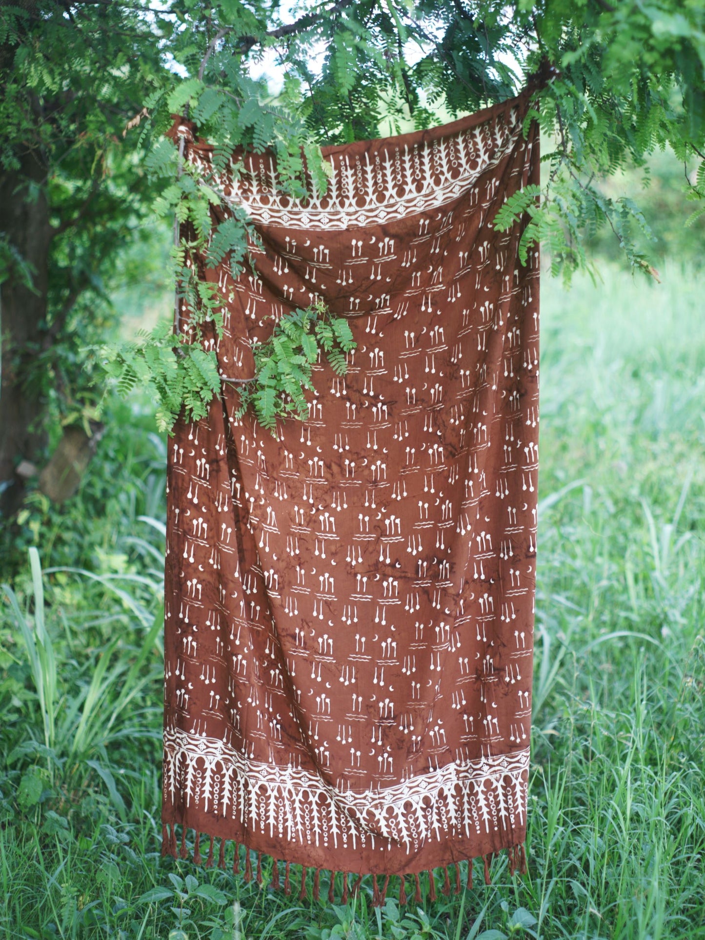 The Hin Kong Bay - Brown sarong from YUMI & KORA hangs from a tree in a meadow. Our sarongs can be used as a home tapestry or as a curtain in your home