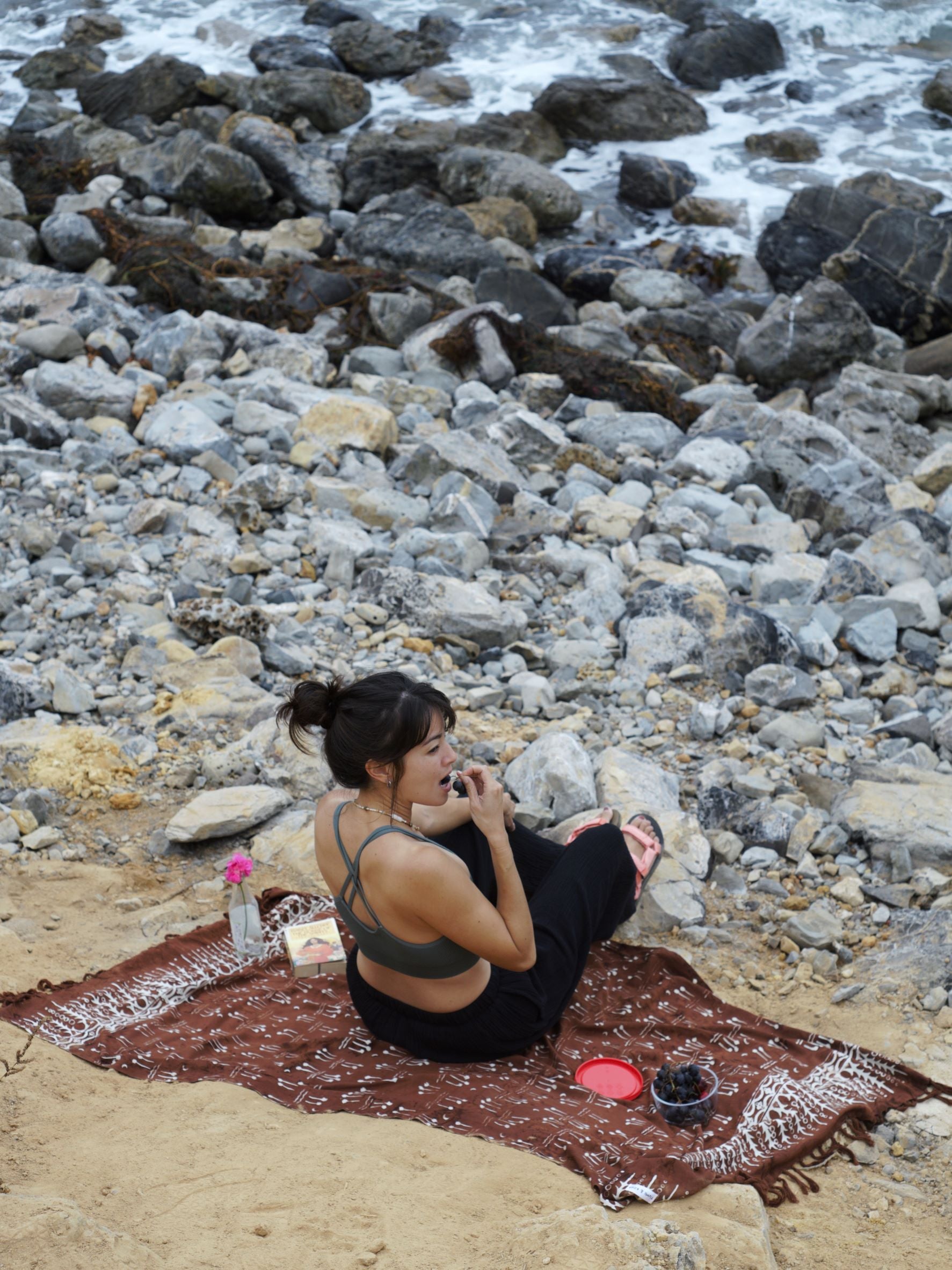 A woman sits on the Hin Kong Bay - Brown sarong from YUMI & KORA by the beach. Our sarongs can be used as a pool towel or as a beach towel