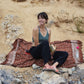 A woman sits on the Hin Kong Bay - Brown sarong from YUMI & KORA by the beach. Our sarongs can be used as a pool towel or as a beach towel