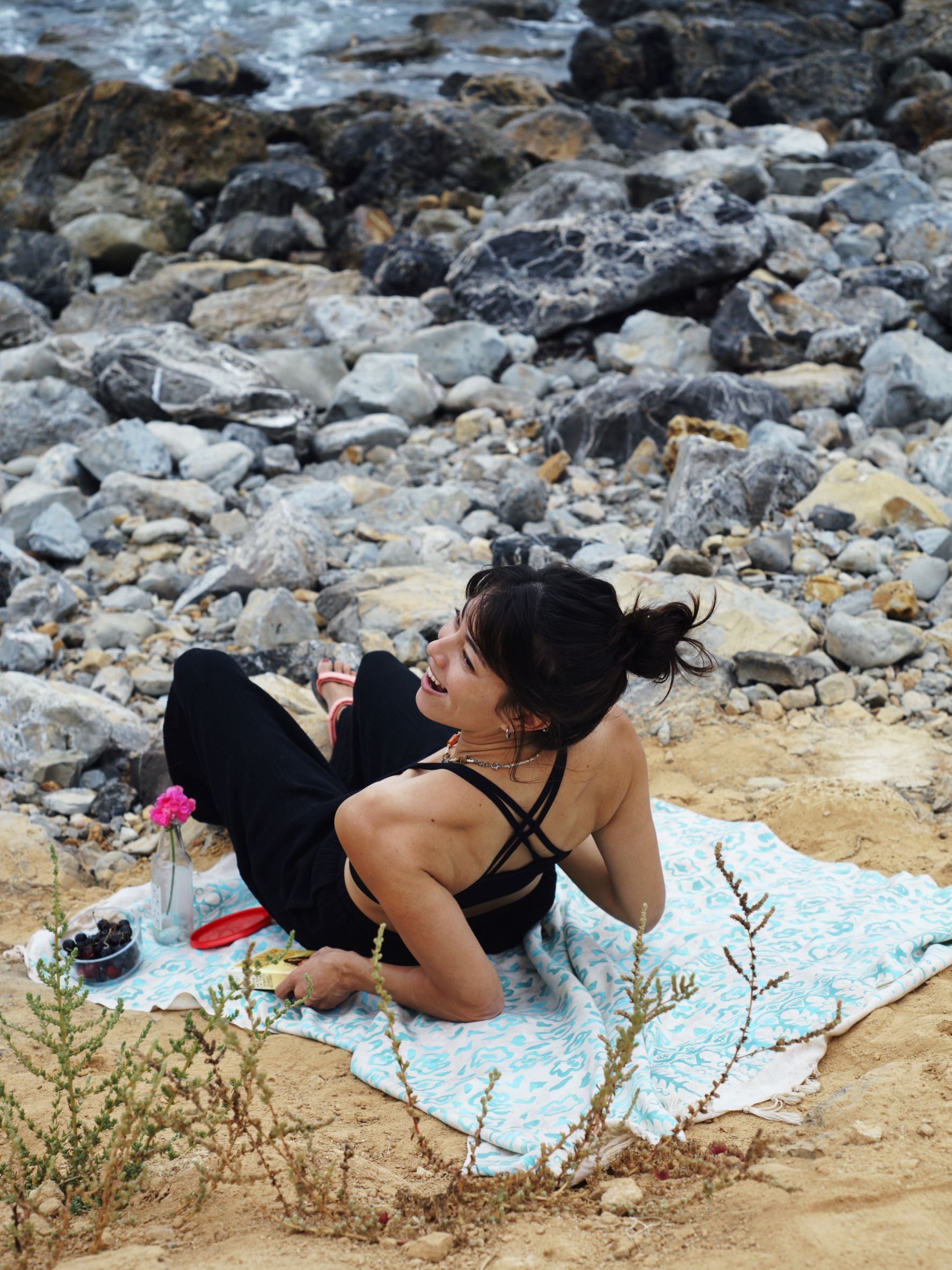 A woman lays on the Summer Bloom - Ivory sarong from YUMI & KORA by the beach. Our sarongs can be used as a picnic blanket or as a beach towel