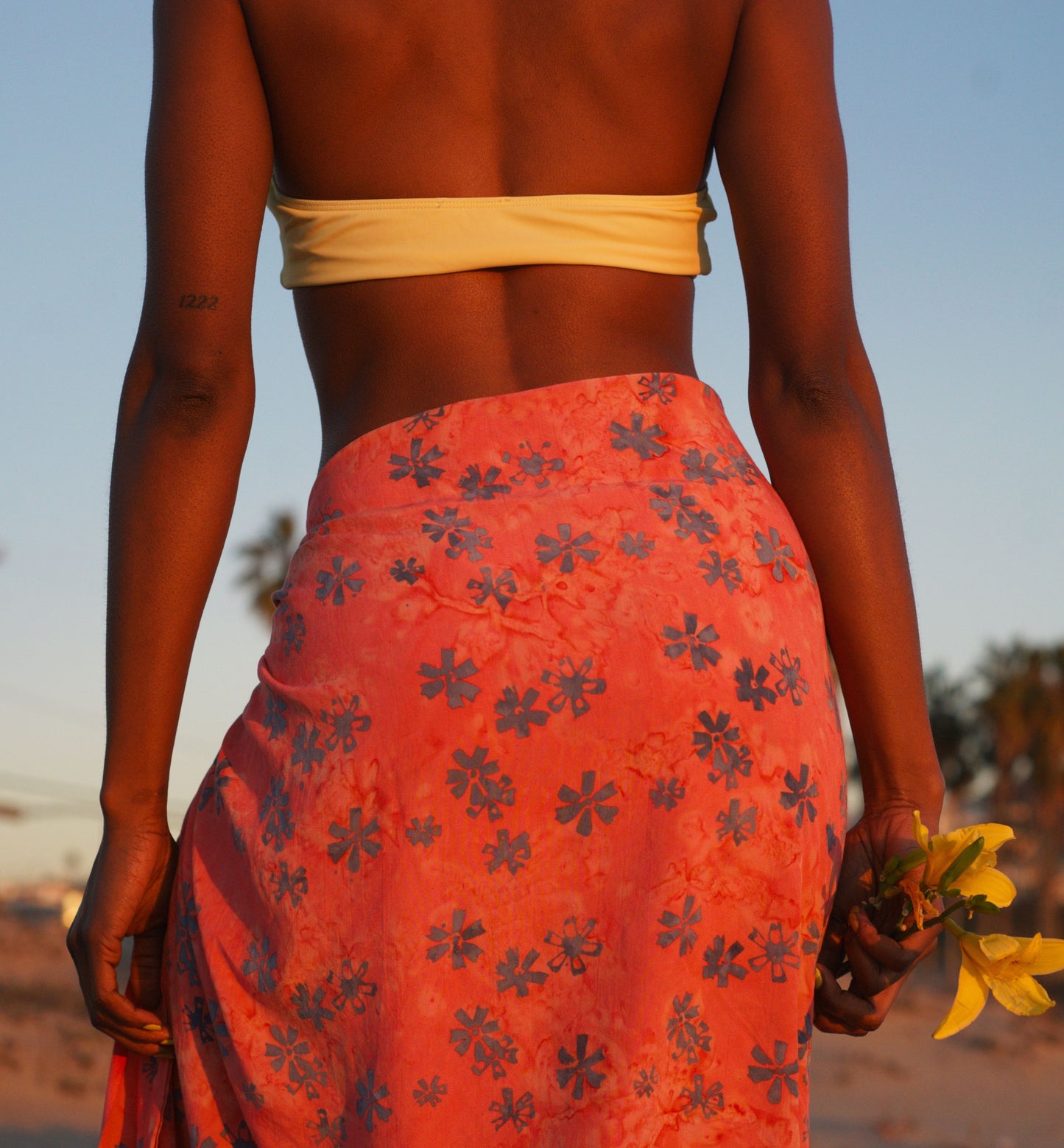 A woman wears the Sweet Cosmos - Pink sarong from YUMI & KORA as a swimsuit cover-up skirt at the beach. Our sarongs can be tied as a skirt, a dress, or a beach kimono