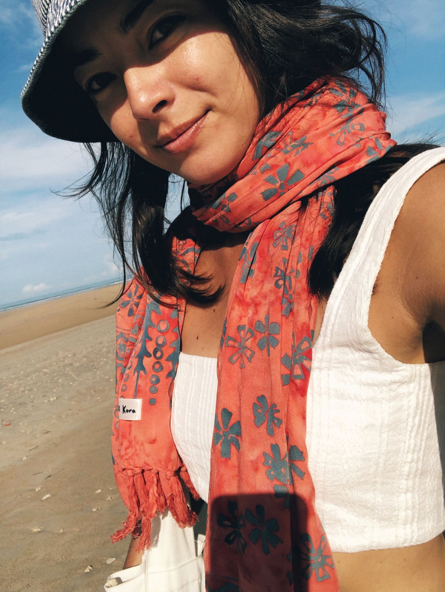 A woman wears the Sweet Cosmos - Pink sarong from YUMI & KORA as a summer scarf, paired with a white crop top and a bucket hat. Our sarongs can be worn as a scarf at the beach