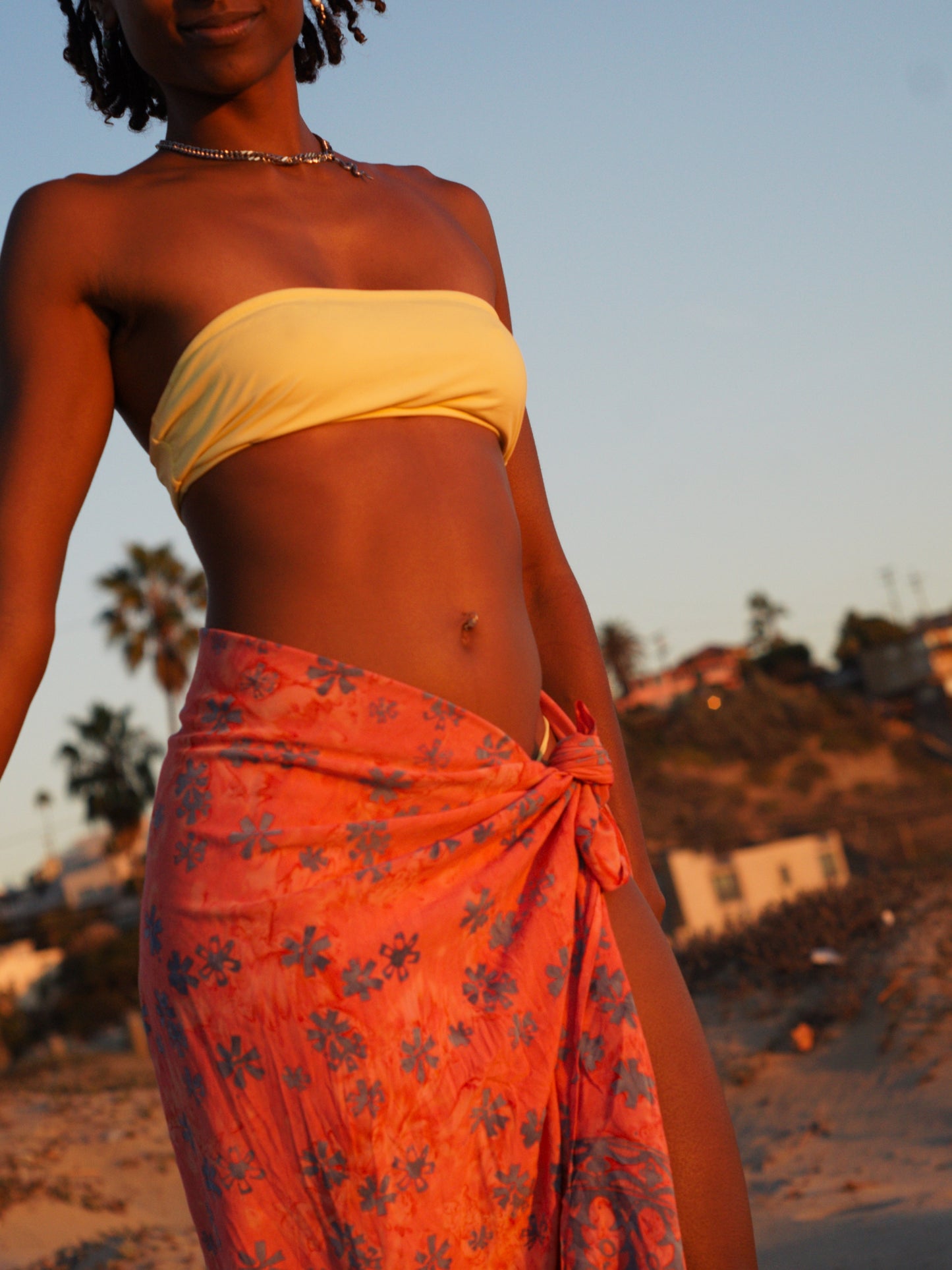 A woman wears the Sweet Cosmos - Pink sarong from YUMI & KORA as a swimsuit cover-up skirt at the beach. Our sarongs can be tied as a skirt, a dress, or a beach kimono